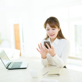 ノートパソコンの前で携帯を操作する女性