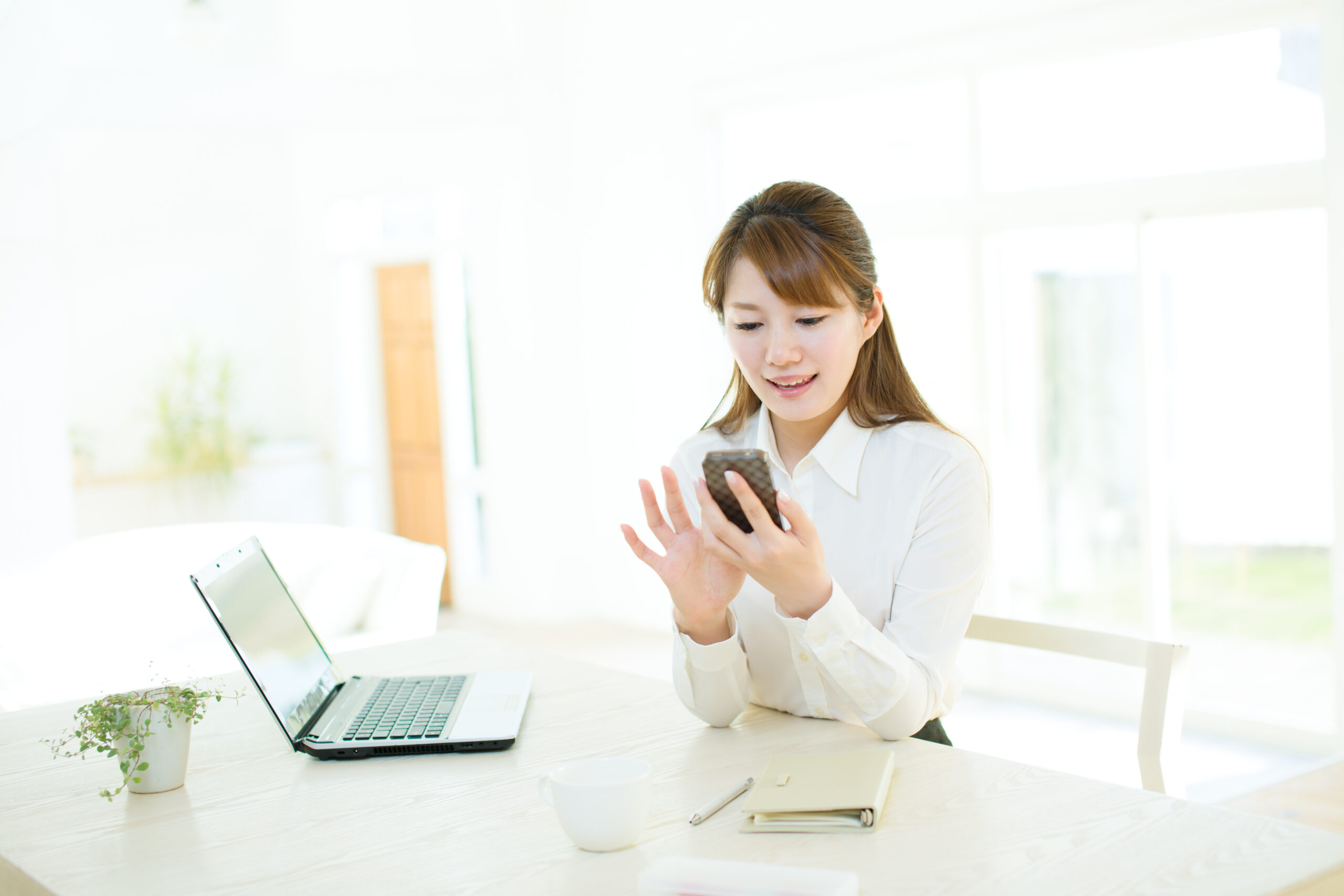ノートパソコンの前で携帯を操作する女性
