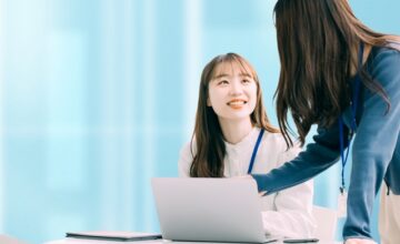 ノートプックパソコンを使っている女性の横に立ってアドバイスをする女性