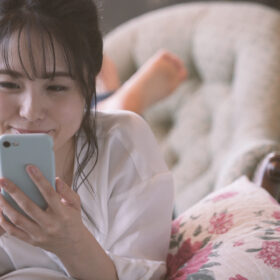 自宅のベットでうつ伏せになってスマホを操作する笑顔の女性